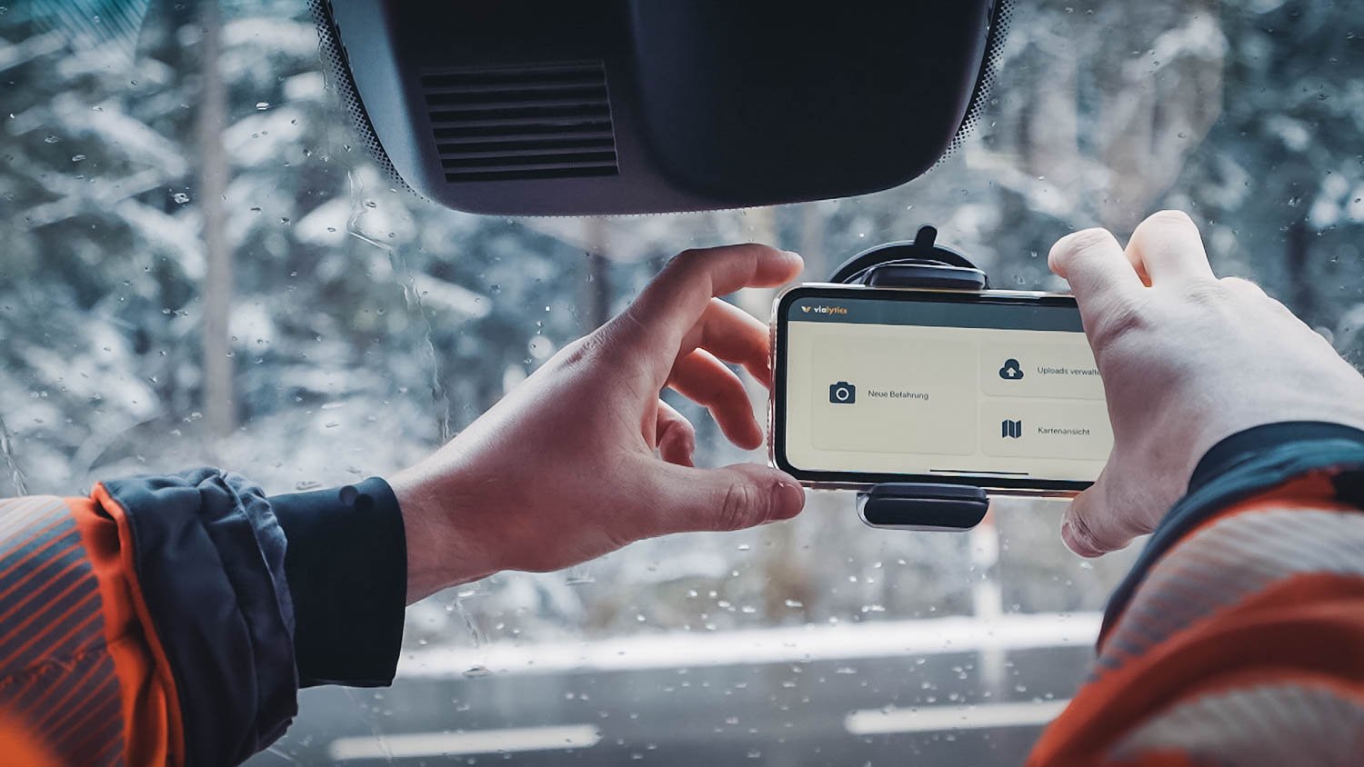 A person from road construction puts a cell phone in a holder on a windshield, in the background a snowy forest can be seen 