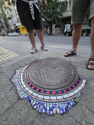 Flacking circulaire encadrant une plaque de regard métallique estampillée « Telefon », dans une rue commerçante d'Ankara. La mosaïque, aux motifs inspirés de la céramique turque (bleu, rouge, blanc, motifs floraux), rayonne autour de la plaque comme un soleil. Deux passants s'arrêtent à côté.