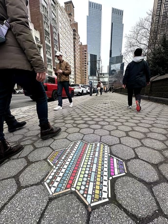 Flacking de forme étoilée incrusté dans un trottoir au dallage hexagonal, dans une rue de New York. La mosaïque multicolore, faite de bandes horizontales de carreaux aux teintes vives, contraste avec le gris de la chaussée usée. Des passants marchent autour.