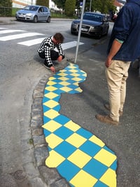 Un homme accroupi sur un trottoir, posant les dernières pièces d'une grande mosaïque à damier bleu et jaune qui s'étend le long d'une bordure en pierre dégradée, près d'un passage piéton. Il porte une veste à carreaux noir et blanc.