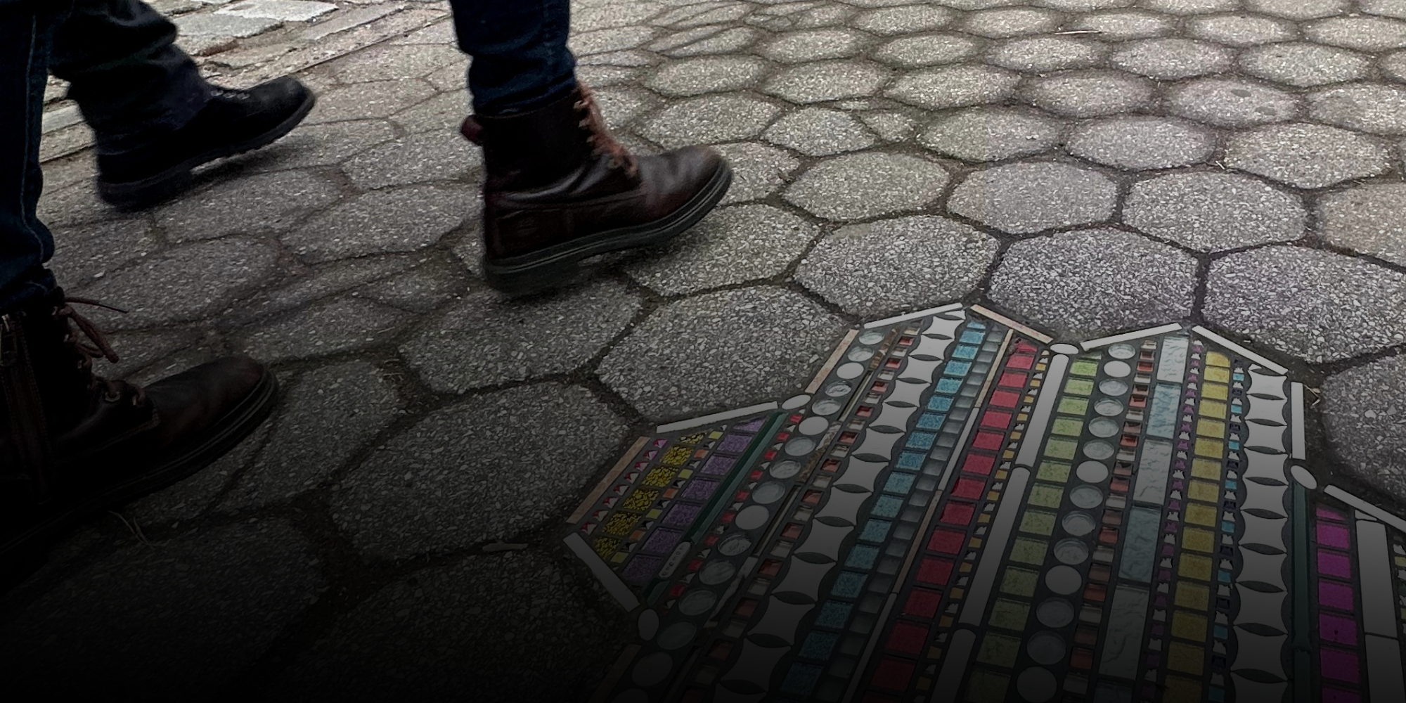 Flacking de forme étoilée incrusté dans un trottoir au dallage hexagonal, dans une rue de New York. La mosaïque multicolore, faite de bandes horizontales de carreaux aux teintes vives, contraste avec le gris de la chaussée usée. Des passants marchent autour.
