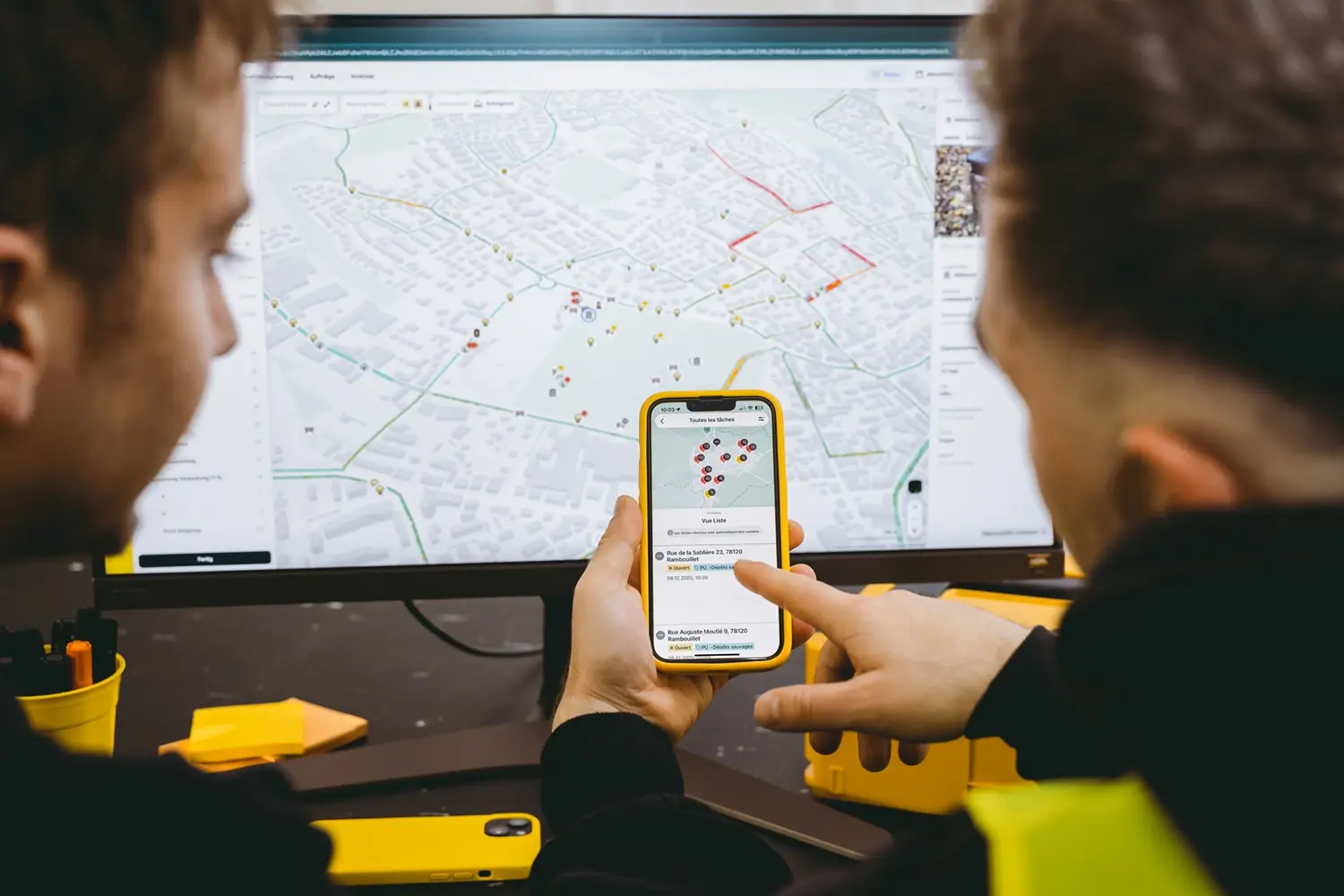 Deux agents en gilet jaune consultent ensemble la plateforme vialytics sur un écran d'ordinateur et un smartphone affichant une carte du réseau routier avec des points de signalement.